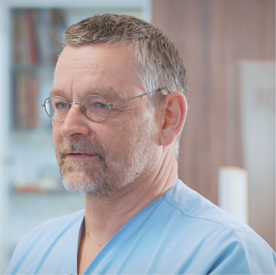 Ein Mann mit Brille und kurzen Haaren trägt ein hellblaues Oberteil und blickt nachdenklich in die Ferne.