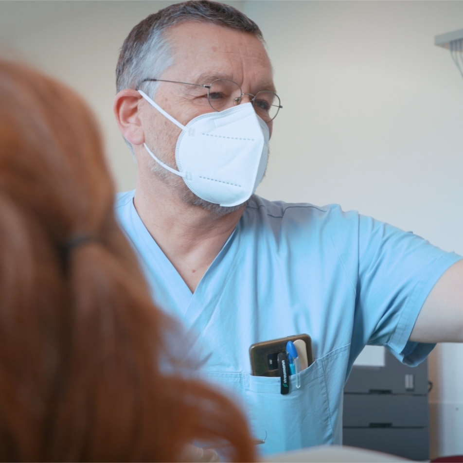 Ein Arzt in blauer Kleidung trägt eine Maske und erklärt eine Behandlung einer Patientin.