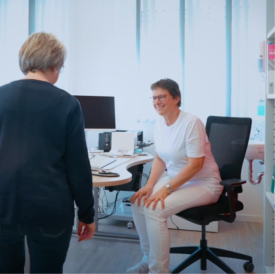 Zwei Frauen in einem hellen Raum, eine sitzt lächelnd auf einem Stuhl, die andere steht ihnen gegenüber.