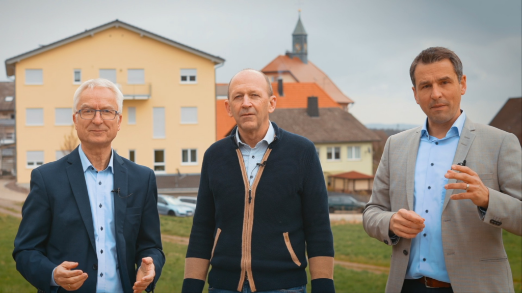 Drei Männer stehen im Freien vor mehreren Gebäuden und sprechen in die Kamera. Der Himmel ist bewölkt.