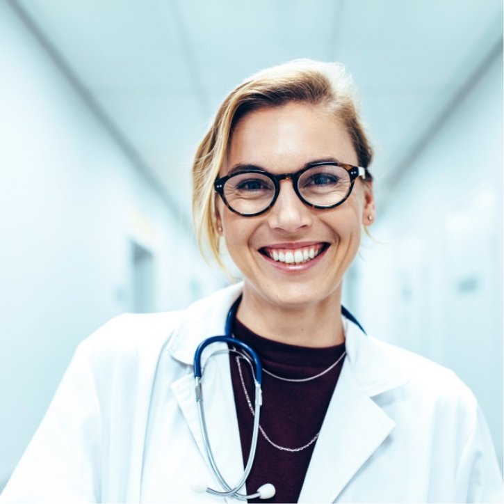 Lächelnde Frau in Arztkittel mit Stethoskop, steht in einem hellen Krankenhausflur.