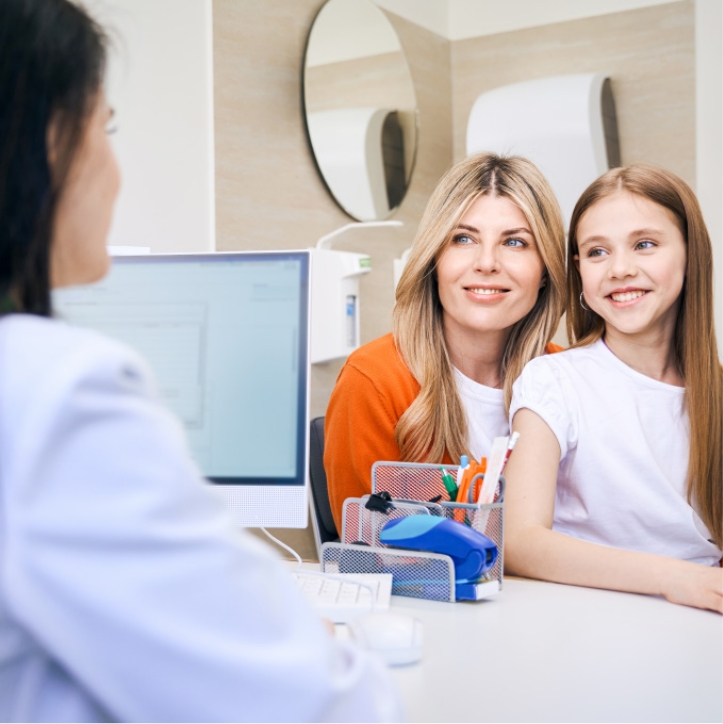 Mutter und Tochter sitzen lächelnd bei einem Arztbesuch gegenüber einer Ärztin am Schreibtisch.